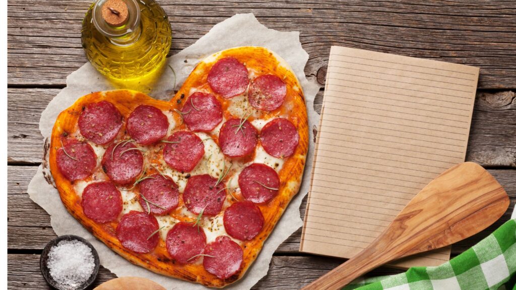 A peperoni pizza in the shape of a heart next to a wooden spoon and notebook.