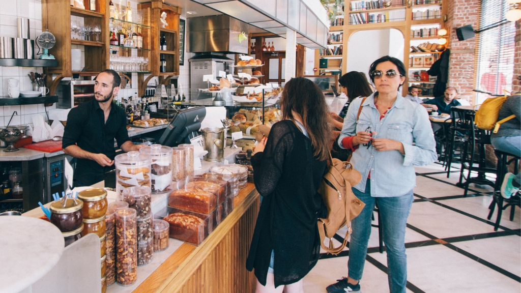 Image is of a busy line waiting to order food at a cafe that serves baked goods, coffee, and custom desserts. A woman in a long black shirt faces the counter to order and another woman in denim faces toward the camera. The shop owner is behind the counter and distracted by incoming patrons. Photo credit: StockSnap via Pixabay